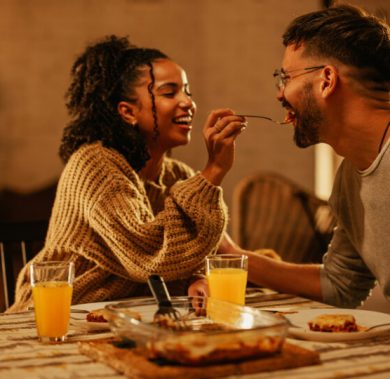 A young and happy couple is having a small dinner at home. The beautiful woman is feeding her boyfriend at the table.